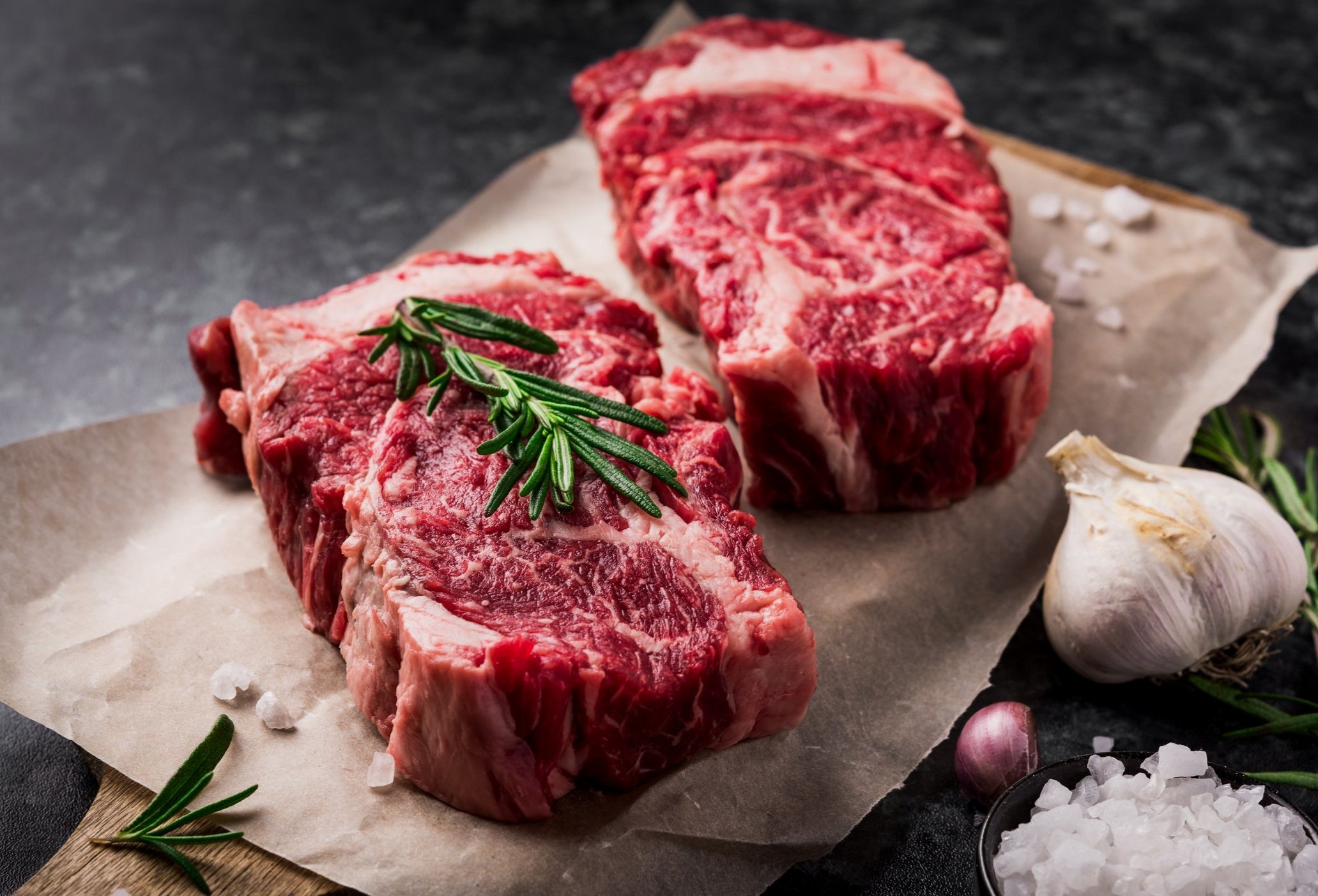 Raw ribeye steaks garnished with fresh rosemary, surrounded by garlic cloves and coarse sea salt on a wooden cutting board. Ideal for grilling or roasting.