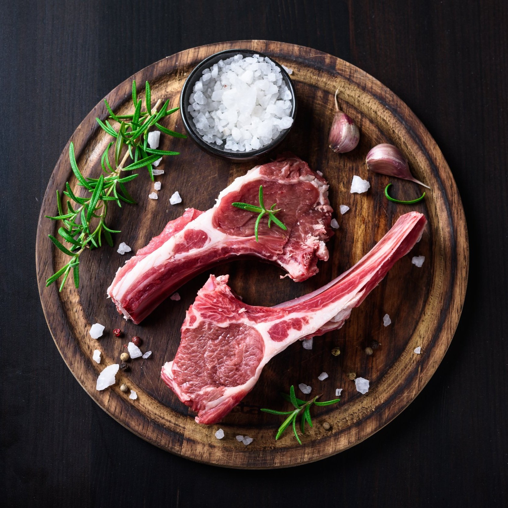 Fresh lamb chops on a wooden platter, garnished with rosemary sprigs and coarse sea salt, accompanied by garlic cloves, ideal for gourmet cooking and grilling recipes.