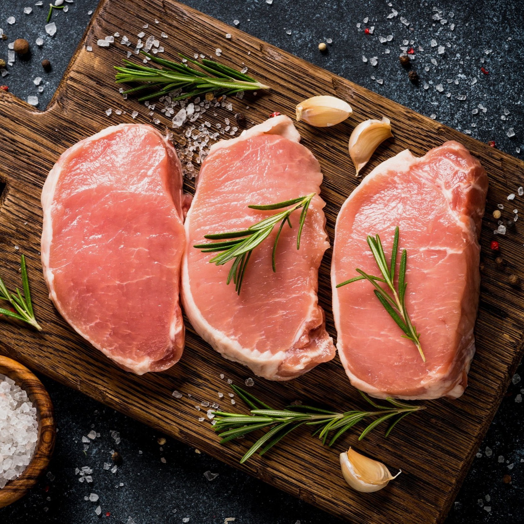 Fresh pork chops garnished with rosemary and garlic, displayed on a wooden cutting board with coarse sea salt and peppercorns. Ideal for cooking enthusiasts and recipe inspiration.
