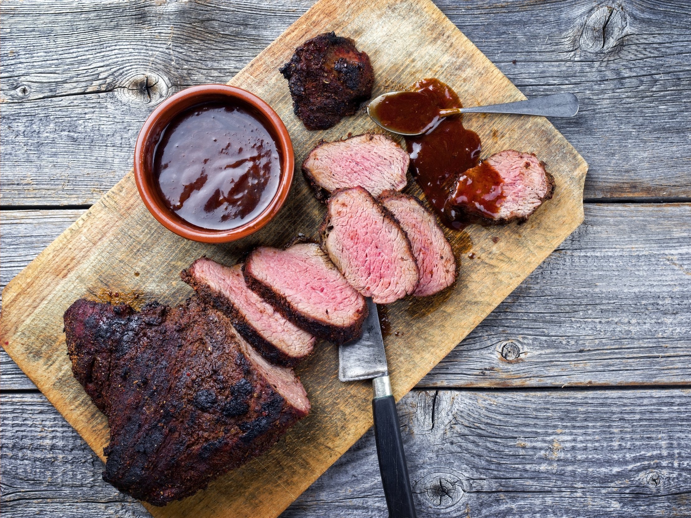 wagyu_tritip_woodboard Sliced smoked beef brisket on a wooden cutting board, accompanied by a small bowl of barbecue sauce and a spoon, showcasing juicy pink meat and a crispy outer crust.